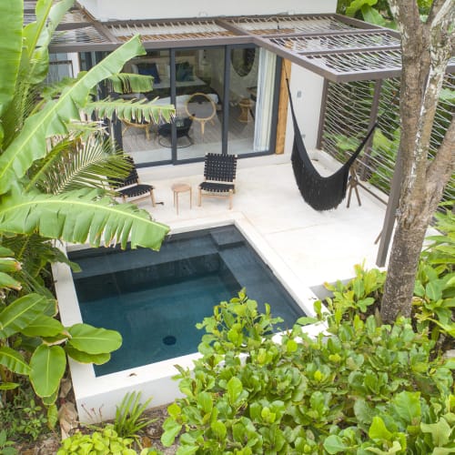 Ein luxuriöser Außenpool ist von üppigen tropischen Pflanzen und Bäumen umgeben, mit einem modernen Haus mit Glastüren und einer Hängematte im Hintergrund.