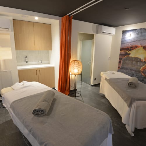 Ein moderner Spa-Behandlungsraum mit zwei Massageliegen, jeweils mit grauem Laken und gerolltem Handtuch, warmem Licht von Stehlampen mit geflochtenen Schirmen und großem naturinspiriertem Wandbild.