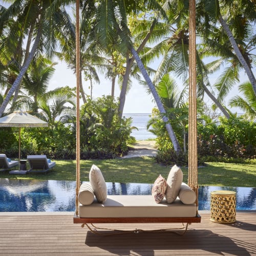 A tropical outdoor scene featuring a hanging cushioned swing with pillows on a wooden deck overlooking a pool, surrounded by palm trees and greenery with a view of the ocean in the background.