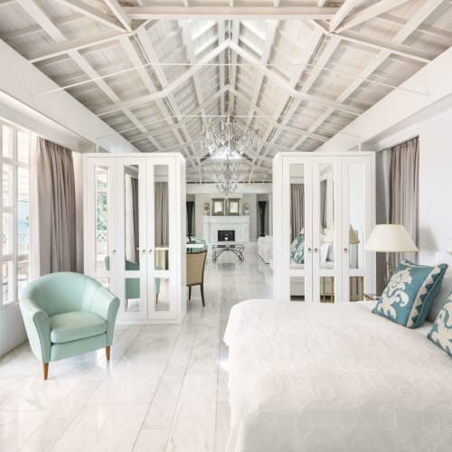 Bright and airy villa interior with white walls, a white bed with patterned pillows, and a light green armchair.