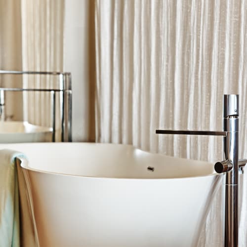 Weiße freistehende Badewanne mit verchromtem Wasserhahn in einem Badezimmer mit beigen Vorhängen und einem hellen Handtuch an der Badewannenrand.