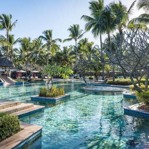 Ein großer Außenpool umgeben von tropischen Palmen und üppigem Grün, mit klarem blauem Himmel und einem Pavillon mit Strohdach im Hintergrund.