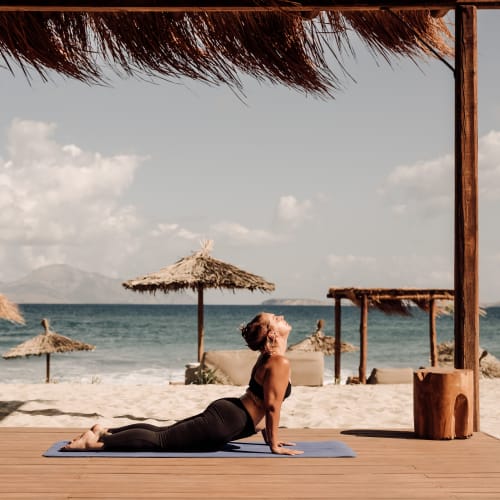 Frau praktiziert Yoga in der Kobra-Pose auf einer blauen Matte unter einem Strohdach am Strand mit Meerblick und bewölktem Himmel.