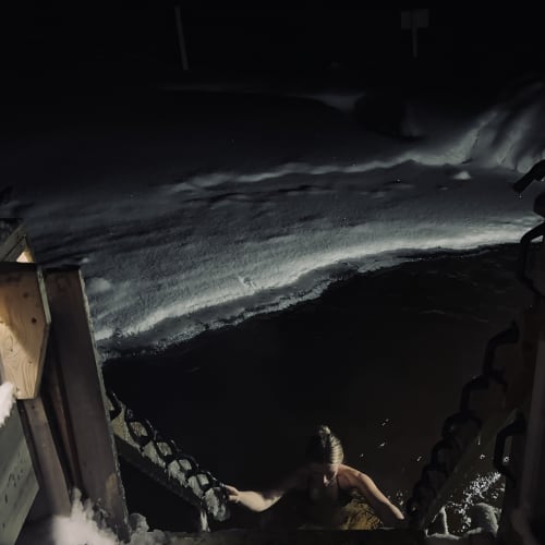 Eine Person klettert bei Nacht aus einem Eisloch in einem zugefrorenen See und hält sich an schwarzen Ketten fest, die mit einer schneebedeckten Plattform verbunden sind.