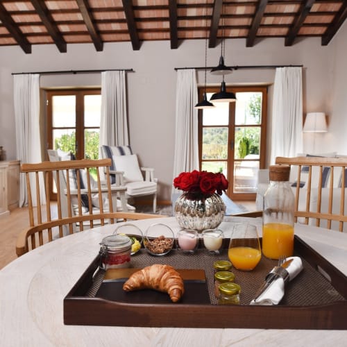 A bright and cozy living area with wooden ceiling beams, white curtains, and a large window overlooking greenery.
