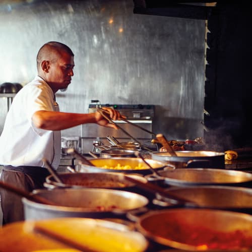 Ein Koch in weißem Hemd kocht mehrere Gerichte in großen Töpfen auf einem Herd in einer professionellen Küche mit metallischer Wand im Hintergrund.