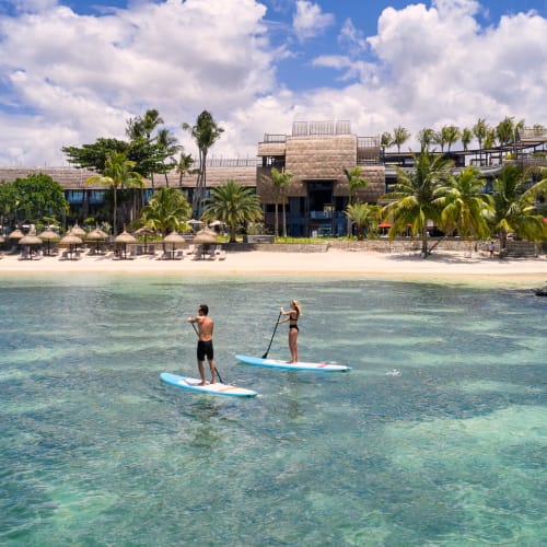 Zwei Personen stehen in der klaren blauen Wassers des Grand Baie, Mauritius, auf Paddleboards mit einem Sandstrand, Palmen und Resort-Gebäuden im Hintergrund.