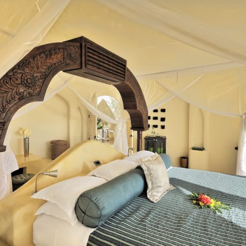 A bedroom in the Kilindi Pavilion in Zanzibar featuring a large bed with white linens and a cylindrical bolster, decorated with a carved wooden arch and surrounded by draped windows and a sea view.