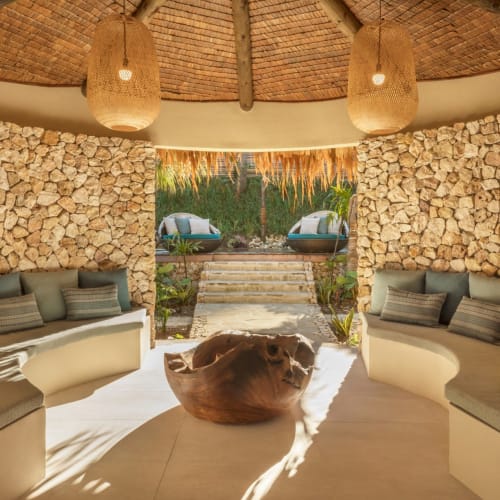 Eine gemütliche, halbkreisförmige Lounge mit Steinenmauern, gepolsterten eingebauten Sitzgelegenheiten und hängenden Korblampen, die in einen Außenbereich mit Stufen zu Stühlen und einem Garten führt.