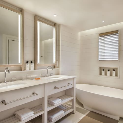 Modern bathroom with double vanity, large mirrors, and a freestanding bathtub under a small window.
