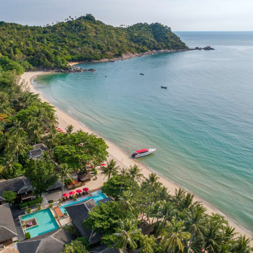 Aerial view of a tropical beach resort with lush greenery, sandy shoreline, and clear blue sea, featuring boats near the shore and resort buildings surrounded by palm trees.