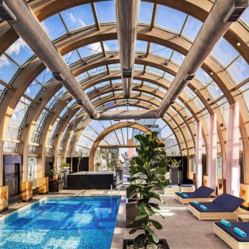 Innenansicht eines luxuriösen Wellnessbereichs mit einem ovalen, gläsernen Gewölbe, Pool auf der linken Seite und sonnenliegen mit Handtüchern auf der rechten Seite.