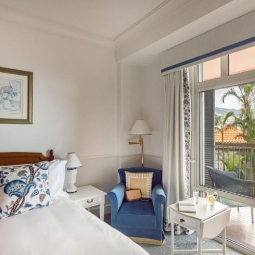 A cozy superior room with a bed, a blue armchair, and a balcony overlooking palm trees and rooftops.