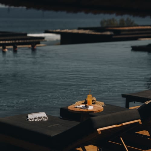 Black cushioned sun loungers with a wooden side table holding a drink, book, and hat beside an infinity pool overlooking the ocean.
