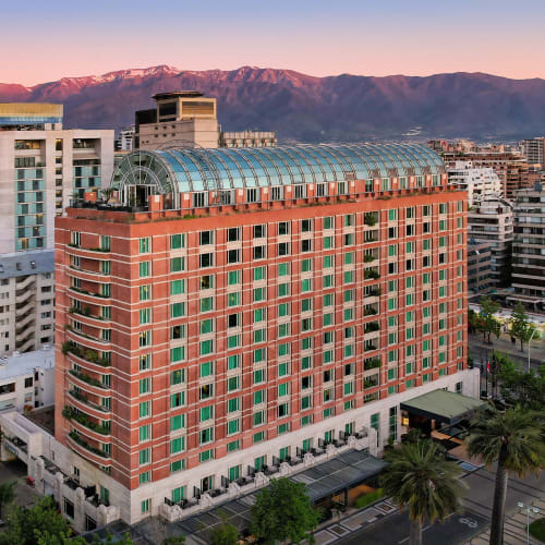 Ein großes, rotes Hochhaus mit vielen Fenstern und einem begrünten Dach in einer Stadt mit Bergen im Hintergrund bei Sonnenuntergang.
