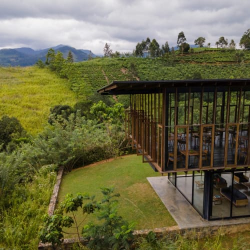 A modern glass-walled building with open-air seating on the upper level, situated on a hillside amidst lush green vegetation and terraced farmland, with mountains in the background.