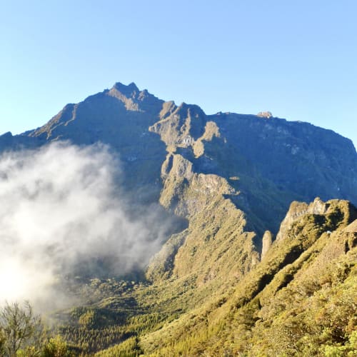 Mountainous landscape with steep rocky cliffs and green forested slopes under a clear blue sky, partly covered by white mist in the valley.