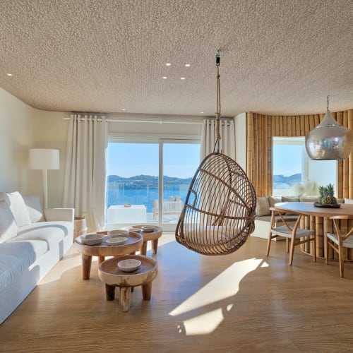 Heller und luftiger Wohnzimmer mit großen Glastüren, die auf einen Wasserblick öffnen, mit weißem Sofa, Holztischen, einem hängenden Korbstuhl und Holzakzenten.