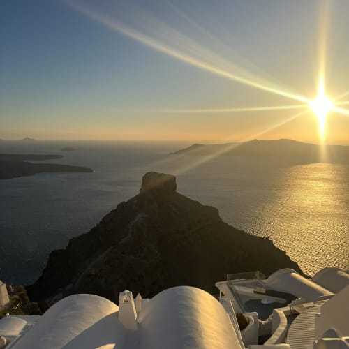 Sun setting over the sea with a silhouetted rocky peninsula and white curved rooftops in Santorini.
