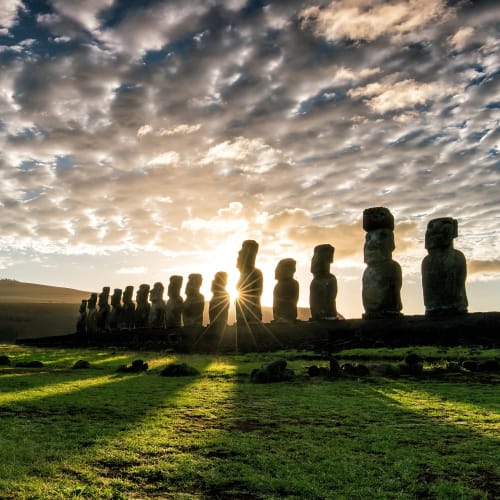 Sunset behind the Moai statues on Easter Island, with clouds in the sky and sun rays peeking between the statues.