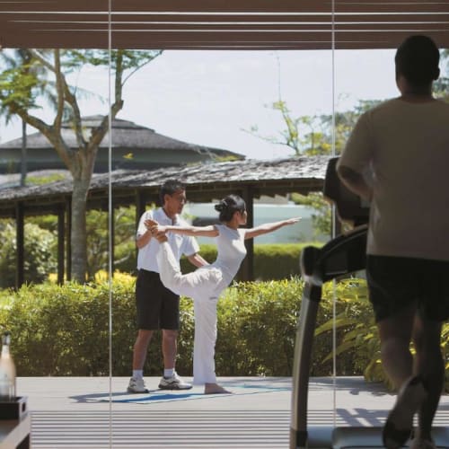 A person jogging on a treadmill inside a gym with large glass windows, observing two children practicing yoga outdoors in a garden.