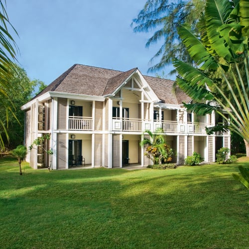 Ein zweistöckiges weißes Holzhaus mit Balkonen im Obergeschoss, umgeben von üppigem grünem Gras und tropischen Bäumen unter klarem blauem Himmel.