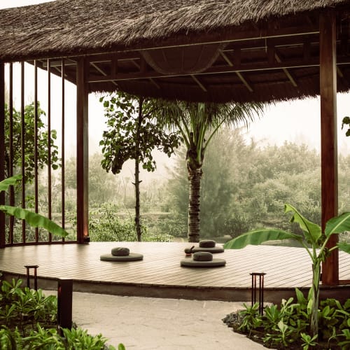 Outdoor meditation sala with wooden floor, cushions, and a thatched roof, surrounded by lush tropical greenery including banana plants and palm trees.
