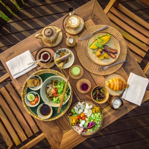 Ein Holztisch mit verschiedenen Gerichten, darunter ein Omelett mit Kartoffeln und Fleisch, eine Salatschüssel, ein Teller mit Frühlingsrollen, ein Croissant sowie eine Teekanne mit Tassen, umgeben von Holzstühlen auf einem Deck.