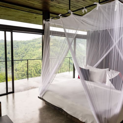 A bedroom with a large bed surrounded by a white mosquito net, overlooking a lush green landscape through floor-to-ceiling glass doors.
