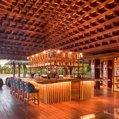 Eine stilvolle Bar aus Holz mit Bambuselementen und warmer Beleuchtung in einem offenen Bereich mit Blick auf Wasser und Palmen in der Ferne.