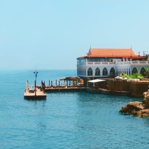 A scenic view of a seaside villa with arches, a terrace, and a boat docked nearby on the calm blue waters.