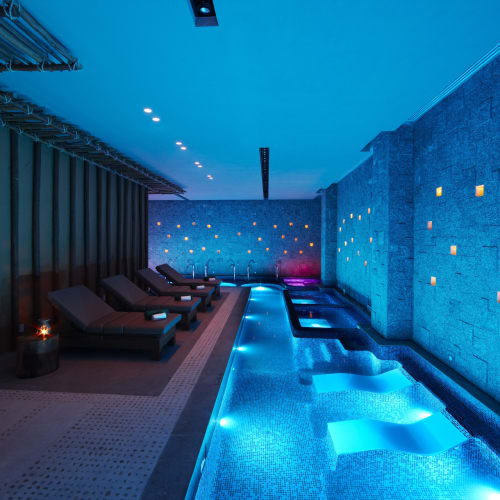 Luxuriöses Spa-Innenbereich mit einem beheizten Pool und mehreren Liegen, beleuchtet mit blauem und warmem gelbem Licht.