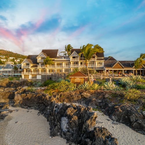 Ein Küstenresort mit großen mehrstöckigen Gebäuden mit Balkonen, umgeben von Palmen und Vegetation, auf felsigem Gelände neben einem Sandstrand bei farbenfrohem Sonnenuntergangshimmel.