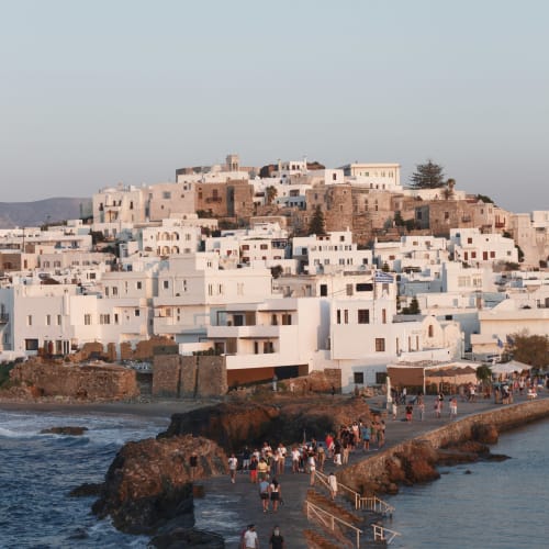 Naxos town with white buildings densely packed on a hillside overlooking the sea, with many people walking along a stone pier extending into the water.