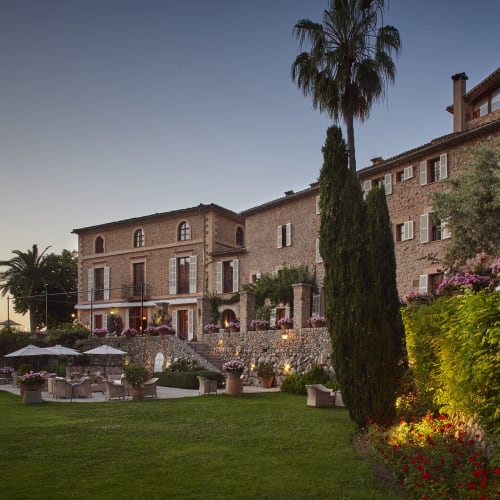 Ein großes historisches Hotel mit Steinwänden und mehreren Fenstern, umgeben von einem gepflegten Garten mit Bäumen, Blumen und Sitzbereichen im Abendlicht.