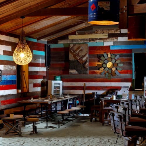 Interior of a rustic bar or café with colorful striped wooden walls, hanging decorative lights, long wooden benches and tables, bar stools, and musical-themed wall decorations including a guitar and a wall sign with positive words.