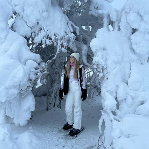 Eine Frau mit weißer Mütze, schwarzen Handschuhen, schwarz-weißer Jacke und weißer Hose steht auf Schneeschuhen in einem verschneiten Wald mit stark frostbedeckten Bäumen.