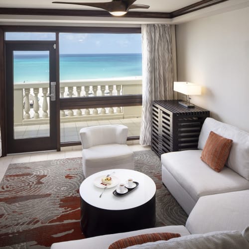 Luxurious hotel room with a white sofa, armchair, and coffee table facing a balcony with ocean view