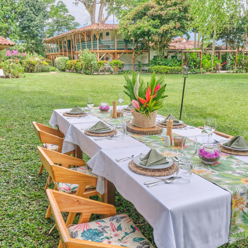 A beautifully set outdoor dining table on a lush green lawn with a tropical table runner and a vibrant floral centerpiece, surrounded by trees and rustic buildings.
