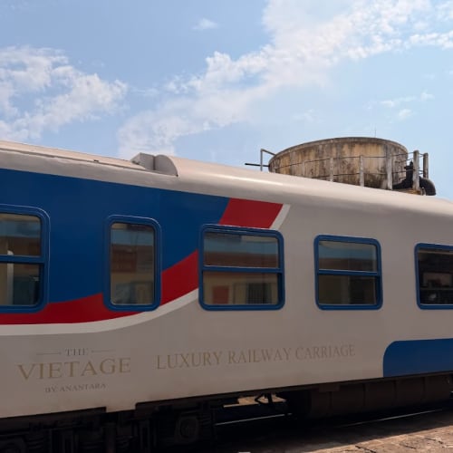 Side view of a vintage luxury railway carriage named 'The Vietage' by Anantara, stationed at a platform under a blue sky with some clouds.