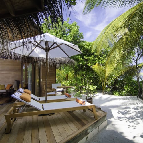 Strandhaus mit Holzdeck, Liegestühlen, Sonnenschirm und üppigem tropischem Grün im Hintergrund.