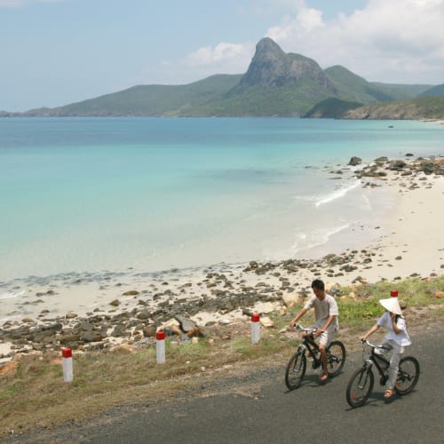 Two people cycle along a coastal road with a beach and mountains in the background.
