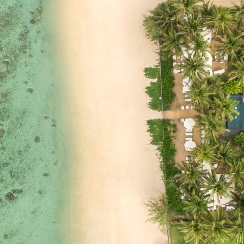 Luftaufnahme eines Strandresorts mit zwei modernen Gebäuden, einem Swimmingpool, umgeben von Palmen, angrenzend an einen Sandstrand und türkisfarbenes Meerwasser.