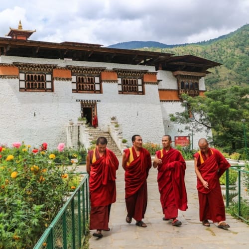 Four monks walking in front of a traditional Bhutanese monastery surrounded by colorful flowers and green hills.