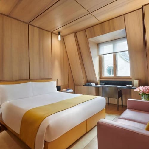 A stylish hotel room with wooden panel walls, a large bed with white bedding and a yellow throw, a pink sofa with decorative cushions, and a window with an angled wall.