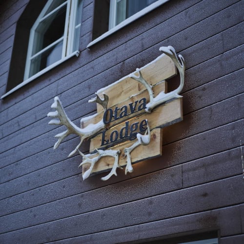 Wooden sign with antlers mounted on a dark wooden exterior wall below two windows reading 'Otava Lodge'.