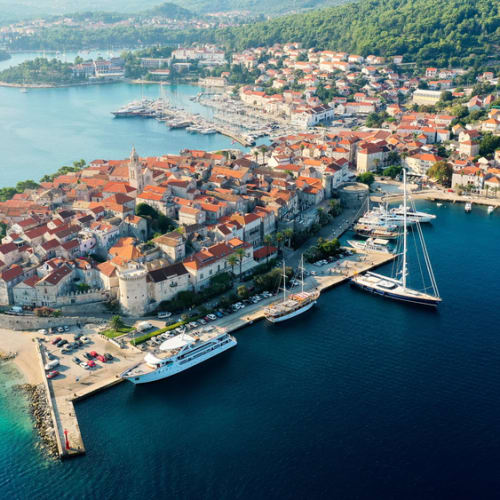 Luftaufnahme der historischen Stadt Korčula mit gewundenen Straßen, roten Dächern und Booten im Hafen, umgeben von grünen Hügeln.