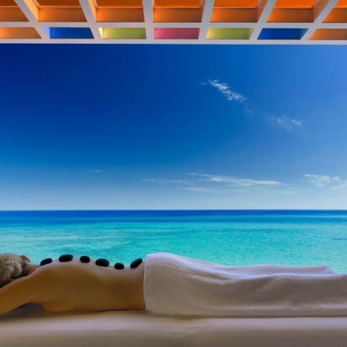 Eine Person liegt auf einer Spa-Liege mit heißen Steinen entlang des Rückens und blickt auf einen schönen Strand mit klarem blauen Wasser und Himmel.