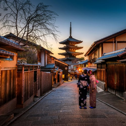 Two women in traditional kimonos walk along a narrow street with wooden buildings towards a pagoda in Kyoto at sunset.
