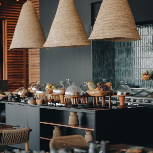 Moderne Küchen- oder Buffet-Anordnung mit hängenden gewebten Pendelleuchten und verschiedenen abgedeckten Gerichten und Obstschalen auf schwarzen Arbeitsplatten.